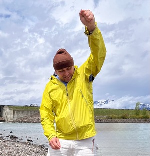 person in a bright yellow raincoat and beanie holding a fish line in front of a lake and forest backdrop
